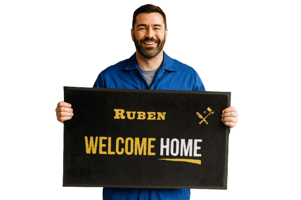 גבר בחולצה כחולה מחזיק בשטיחון דלת שחור שעליו כתוב "RUBEN WELCOME HOME" באותיות צהובות ולבנות — השראה מושלמת לשטיחי כניסה לעסקים ולבניינים.